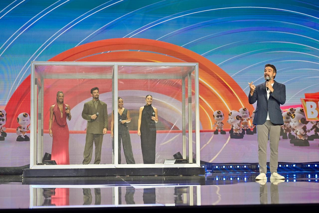 Os ex-BBBs Aline, Vinícius, Pitel e Renata simulam estar dentro de uma casa de vidro; do lado de fora da casa está Tadeu Schmidt gesticulando e segurando um microfone.