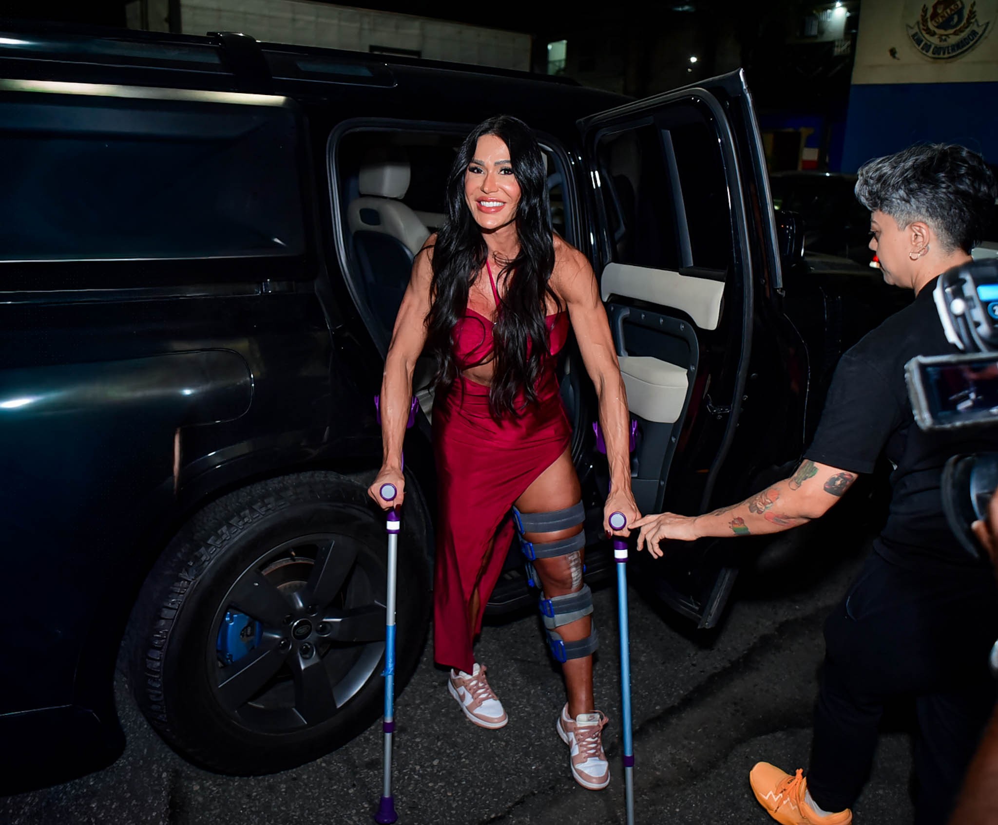Gracyanne Barbosa saindo de um carro, usando um vestido vermelho e a perna numa tala azul, se apoiando em muletas com a ajuda de outra pessoa.