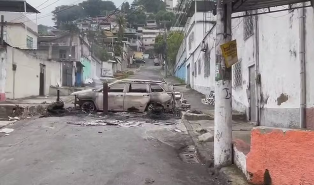 Operação Contenção mobiliza 2,5 mil agentes contra o crime organizado no RJ um carro queimado durante uma barricada, no fundo uma comunidade