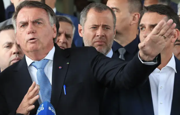 Homem de terno faz gesto com o braço estendido enquanto fala diante de microfones, cercado por outros homens em ambiente público.