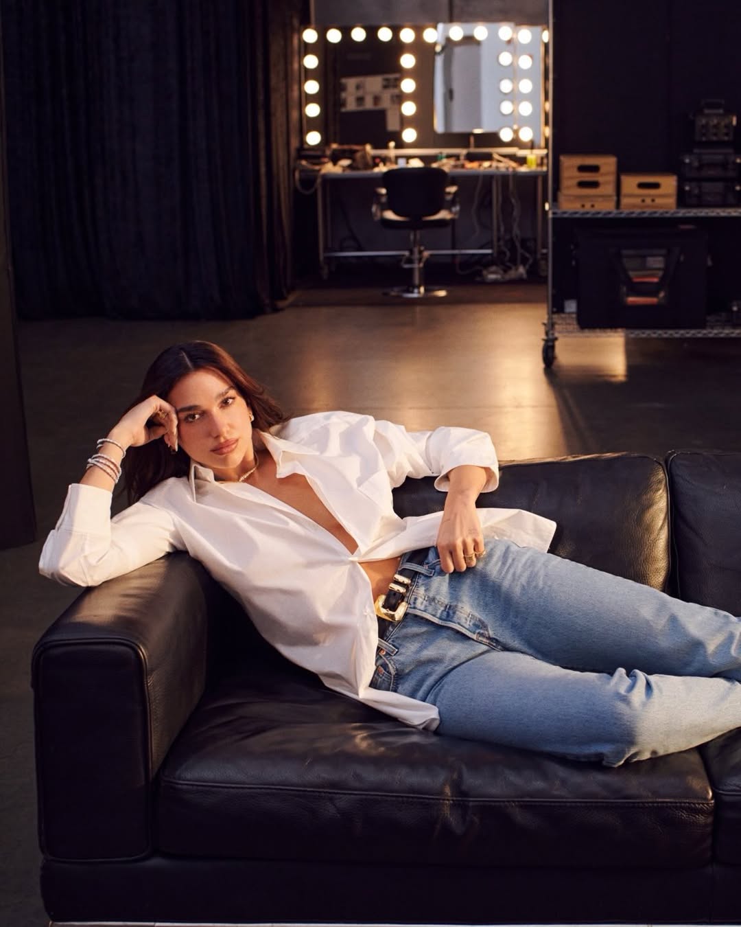 Dua Lipa é escolhida como nova embaixadora global da Bvlgari Dua lipa é uma mulher branca com cabelos e olhos castanhos, ela está deitada em um sofá usando uma camisa branca e uma calça jeans.