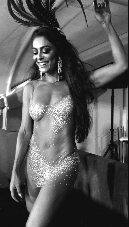Juliana Paes transforma vestido de 2018 em hit fashion do Carnaval 2026 Imagem em preto e branco de uma mulher posando em ambiente interno, com clima de bastidor ou camarim. Ela está de pé, com o corpo levemente inclinado para um dos lados, sorrindo de forma espontânea enquanto olha para baixo, como se estivesse em um momento descontraído.Tem cabelos longos e escuros, soltos sobre os ombros e pelas costas. Na cabeça, usa um adereço alto com penas escuras que se projetam para cima, criando uma silhueta dramática e elegante. Os brincos são longos e brilhantes, pendendo até perto do pescoço. Ela veste um figurino transparente e justo ao corpo, coberto por pequenas pedrarias ou cristais que formam desenhos e brilham mesmo na imagem sem cor. A peça é sem mangas, com alças finas, marcando a silhueta e deixando pernas e braços à mostra. O brilho das aplicações cria contraste com o fundo mais escuro. Um dos braços está levantado, segurando ou ajustando o adereço de penas, enquanto o outro permanece relaxado ao lado do corpo. Ao fundo, aparecem elementos de bastidores, como uma parede clara e estruturas que lembram suporte de iluminação ou equipamentos de palco. A imagem transmite elegância, confiança e leveza, captando um instante íntimo antes ou depois de uma apresentação festiva.