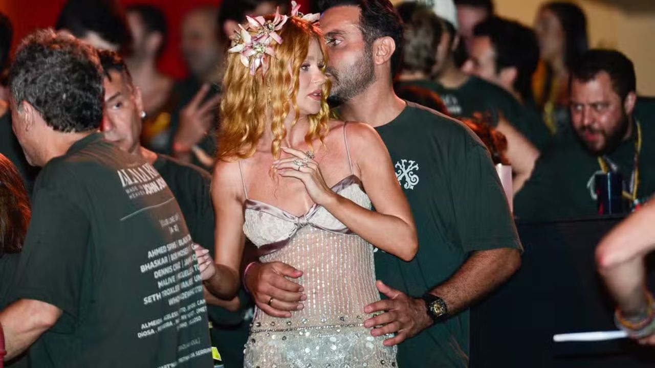 A mulher tem pele clara e cabelos longos, ondulados, em tom ruivo intenso. Ela usa um vestido claro, ajustado ao corpo, com parte superior estruturada semelhante a um corset e detalhes brilhantes na saia. Na cabeça, usa um acessório com flores em tons suaves, que combina com o visual delicado. Sua expressão é séria e ela olha para o lado, com uma das mãos próxima ao peito. O homem está atrás dela, abraçando-a pela cintura com as duas mãos. Ele tem pele clara, cabelos escuros curtos e barba aparada. Veste uma camiseta verde escura e um relógio no pulso. Seu rosto está próximo ao dela, como se estivesse falando algo ao seu ouvido.