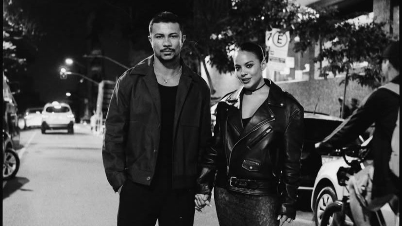 A imagem em preto e branco mostra Bella Campos e Xamã caminhando juntos em uma rua à noite. Os dois estão lado a lado, de mãos dadas, ocupando o centro da cena. Xamã aparece à esquerda, com expressão séria e olhar voltado para a câmera. Ele veste uma jaqueta escura sobre uma camiseta preta, com um estilo casual e urbano. Sua postura é firme e tranquila. À direita, Bella Campos olha diretamente para a câmera com um leve sorriso. Ela usa uma jaqueta de couro escura, ajustada ao corpo, e um acessório no pescoço, o que dá um visual mais moderno e marcante. Seu cabelo está preso, destacando o rosto. O cenário ao redor mostra uma rua com carros estacionados e algumas luzes ao fundo, indicando ambiente urbano. Há também uma pessoa ao lado direito da imagem, parcialmente visível, segurando uma bicicleta. A iluminação vem de postes e luzes da rua, criando contrastes fortes típicos de fotos noturnas em preto e branco.