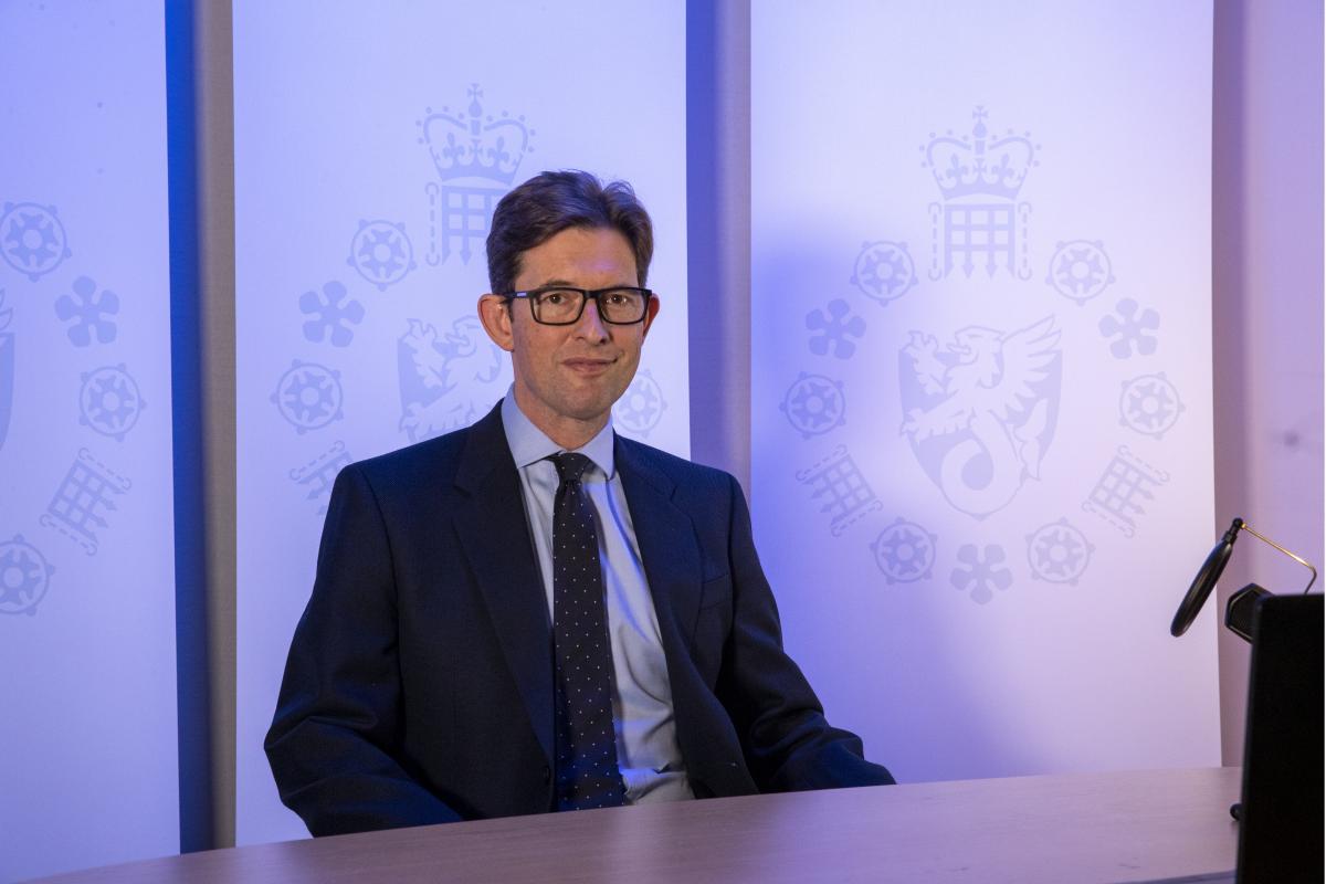O diretor do MI5, ken maccalum. ele usa um terno azul marinho com uma camisa azul clara e gravata preta com pontinhos. atrás dele, um fundo azul com o distintivo do mi5