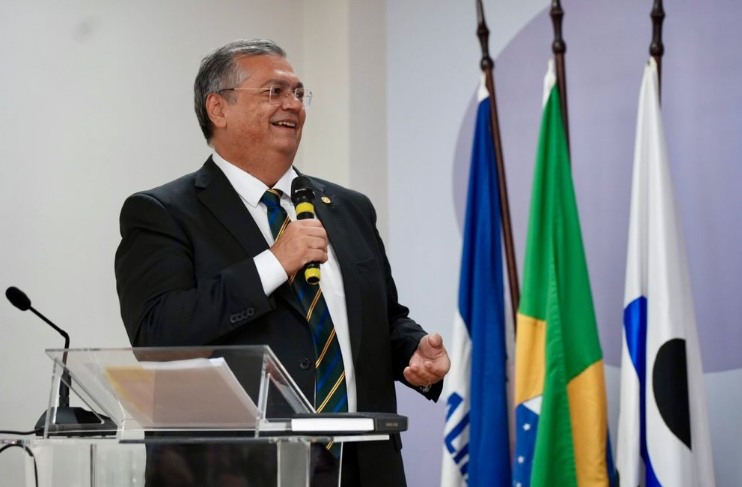 A imagem mostra Flávio Dino em um ambiente interno, de pé e falando ao microfone. Ele é um homem de meia-idade, com cabelos curtos e grisalhos e óculos de armação fina. Ele está sorrindo levemente enquanto olha para fora da moldura, para a esquerda, e segura um microfone de mão em sua mão direita perto de sua boca. Ele está vestindo um paletó preto sobre uma camisa branca de botões e uma gravata listrada com listras de azul, verde, branco e amarelo. Atrás dele, há um fundo desfocado. Três bandeiras nacionais estão dispostas atrás e à direita dele, em mastros. Da esquerda para a direita: uma bandeira azul e branca (bandeira do estado do Maranhão), uma bandeira verde e amarela (bandeira do Brasil) e uma bandeira branca com um círculo preto sobre ela (bandeira do município de São Luís).