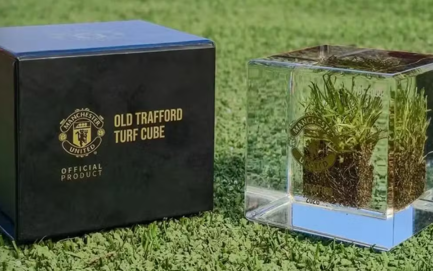 Caixa preta e cubo de acrílico transparente do Manchester United apoiados sobre um gramado verde. O cubo de acrílico contém uma amostra de terra com grama preservada em seu interior, e a caixa preta exibe o escudo e nome do time em dourado.