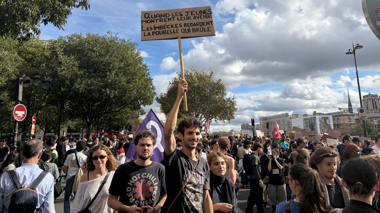 Paris vira zona de guerra em meio a manifestações um jovem francês segura um cartaz chamando os jovens para protestar. em volta deles, várias pessoas.